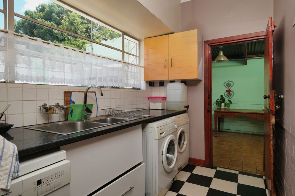 A single white front-loading machine built into a narrow kitchen run beside the sink and dishwasher, with a curtained window above and a chequerboard tiled floor