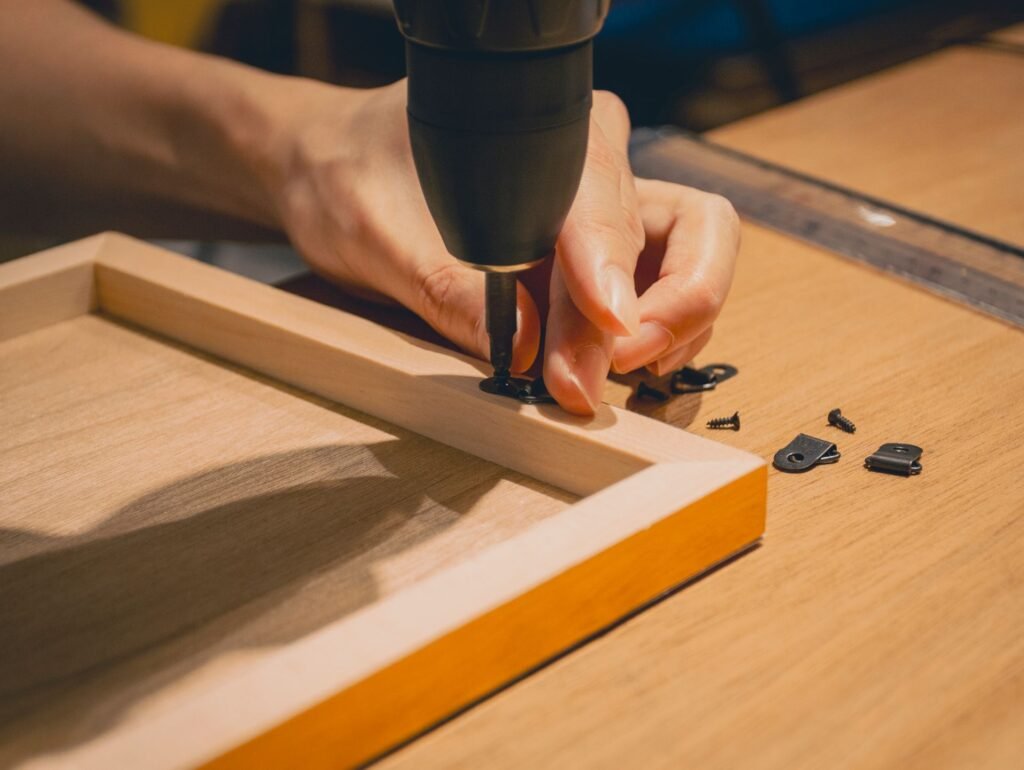 Hands using a cordless driver to fasten a small black metal bracket and screws to a timber frame on a workbench