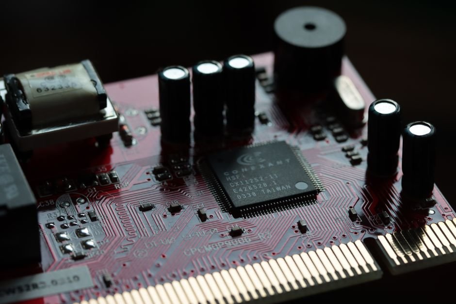 A dark red printed circuit board lit from one side, showing a square controller chip, a row of black cylindrical capacitors, a transformer and a gold edge connector, the layout typical of a washer inverter board