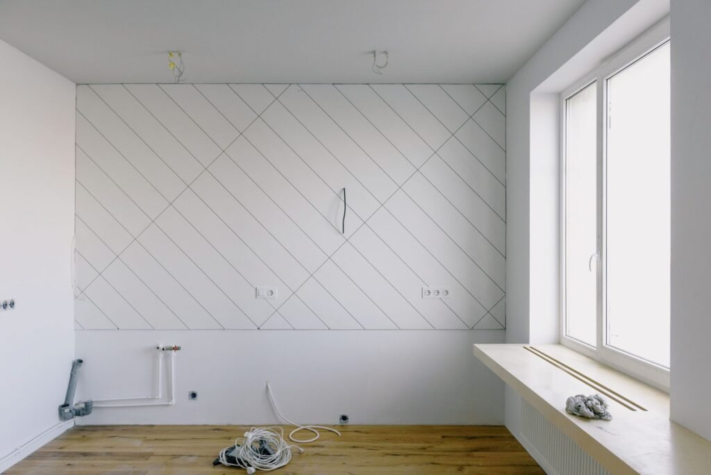 An empty room mid-renovation with capped water supply stubs and a shutoff manifold low on one wall beside a grey waste trap stub and unterminated electrical boxes, the rough-in stage at which a washer outlet box is set