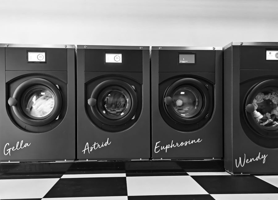 Four front-load washing machines in a row in a black and white photograph, each with a name painted on the cabinet, standing on a chequerboard tiled floor