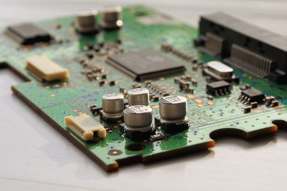 A green printed circuit board photographed at a low angle on a white surface, showing five silver electrolytic capacitors, a square microprocessor and two pale ribbon connectors of the kind a washing machine control board uses
