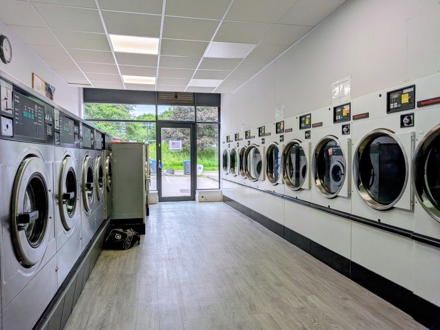 A laundromat with two facing rows of front-load washing machines, their round chrome-rimmed doors and handles clearly visible along a pale wood-effect floor leading to a glass entrance door