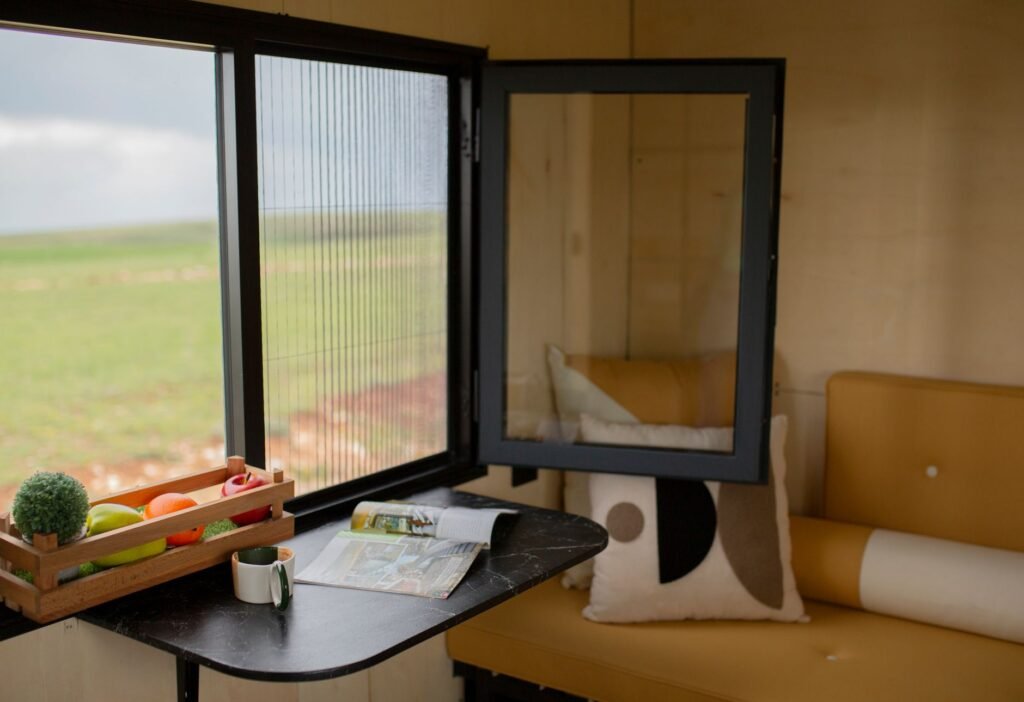 Tiny house interior with plywood walls, a fold-down black table and a mustard bench seat beside an open window onto grassland, the kind of footprint a washing machine for tiny house use has to share