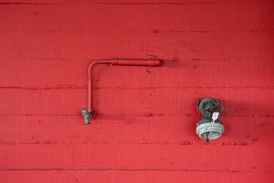 A small water tap on a short pipe and a capped supply fitting mounted on a red painted wall, the kind of exposed supply connection a washing machine leak detector is fitted to