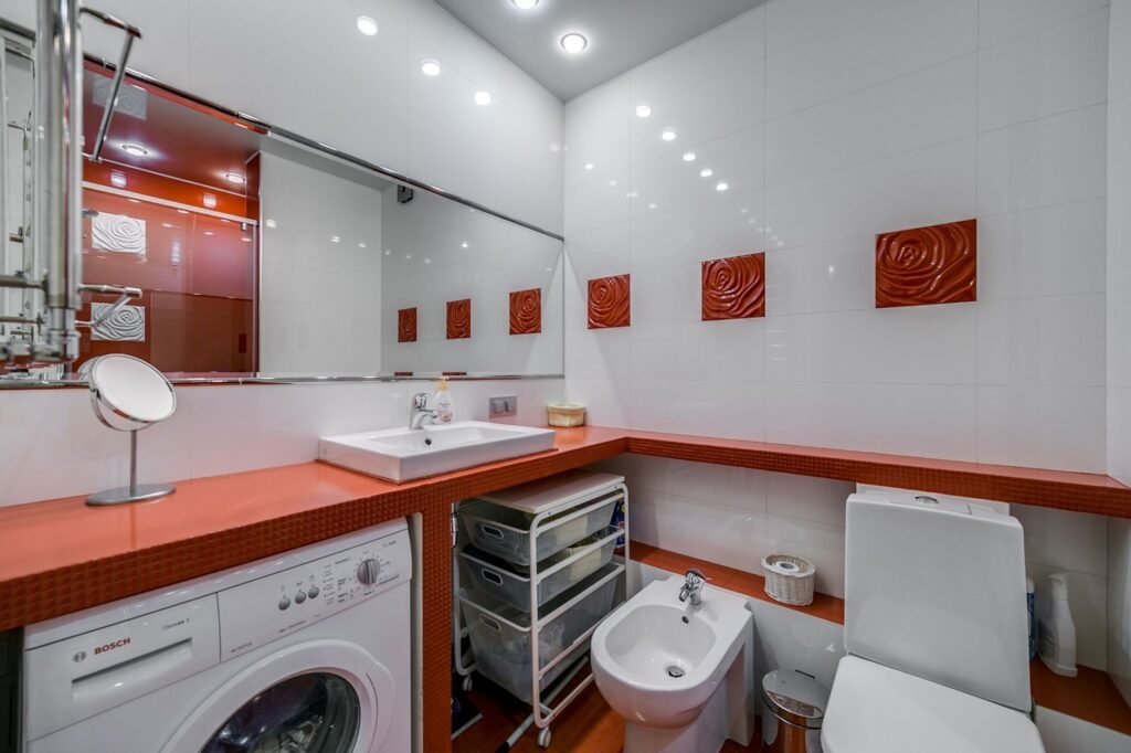 A white Bosch front-load washing machine built in beneath an orange worktop in a small white-tiled bathroom, with a sink, mirror and storage trolley alongside