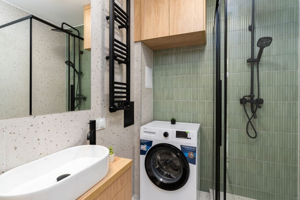 A front-loading washing machine squeezed between a basin unit and a glass shower in a narrow flat bathroom