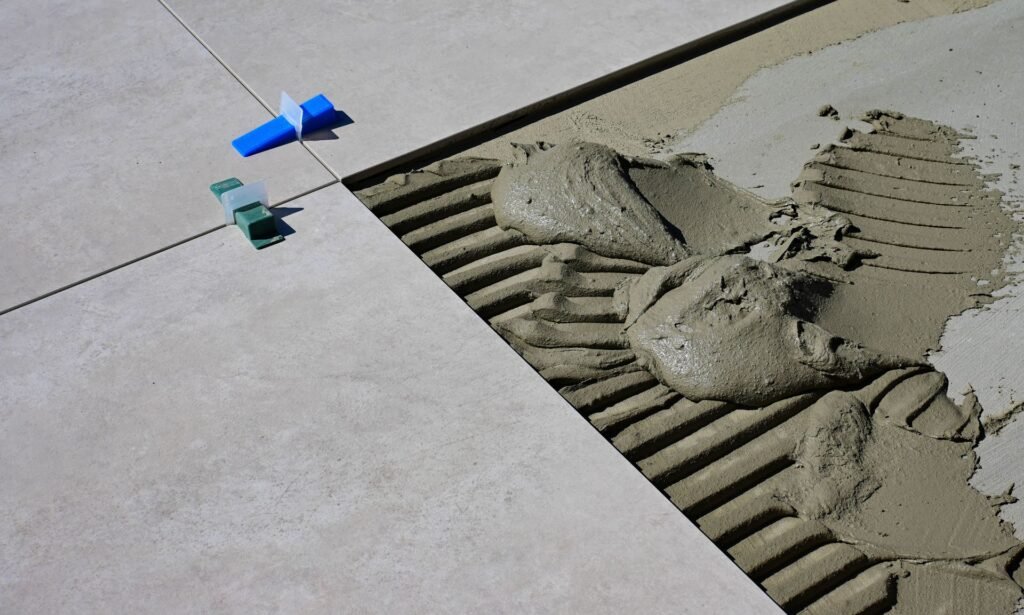 A large pale floor tile bedded into combed grey thin-set mortar with blue levelling clips in the joint, the layer under the surface that does the real work when waterproofing a laundry room floor
