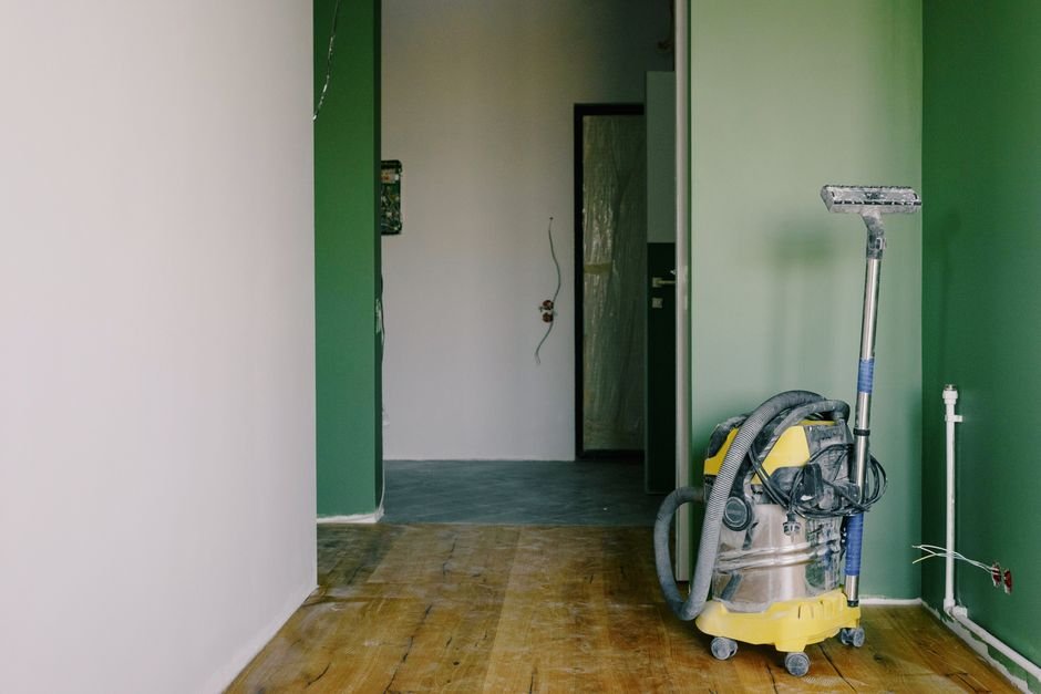 Yellow and stainless wet dry vacuum with its hose coiled over the drum and a floor wand clipped on, standing on bare floorboards in a half-painted room