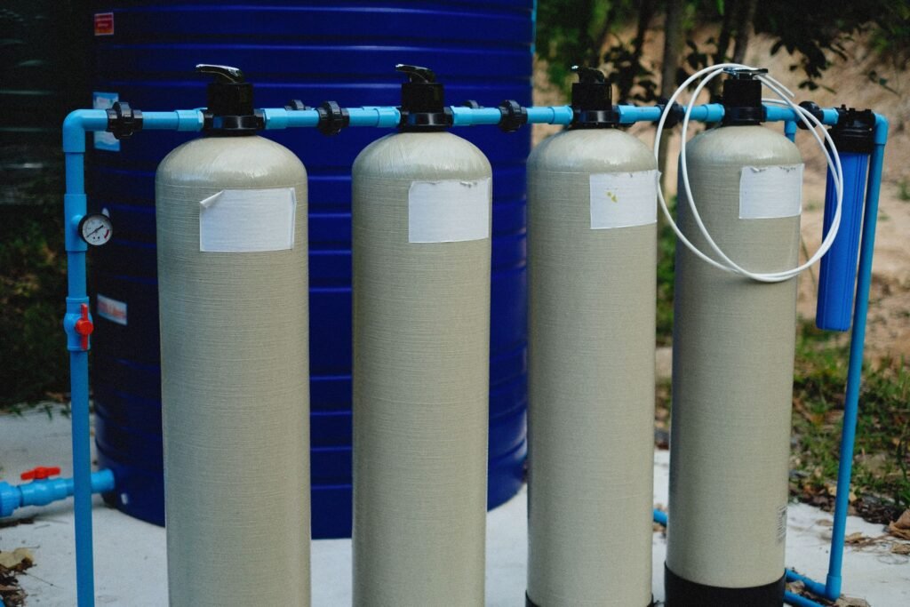 Four fibreglass water treatment tanks on a blue PVC manifold with a pressure gauge and a cartridge housing
