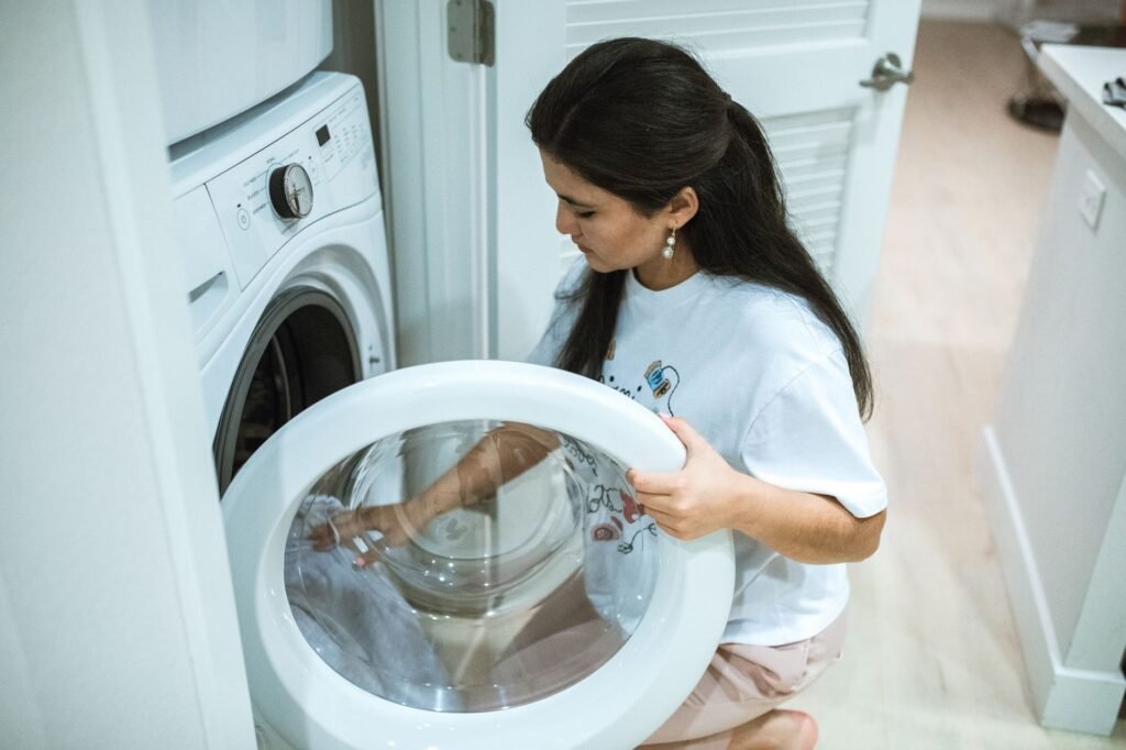 A woman kneeling at a stacked laundry pair, holding the glass door of the lower machine open and reaching into the drum, the moment a sheet goes in