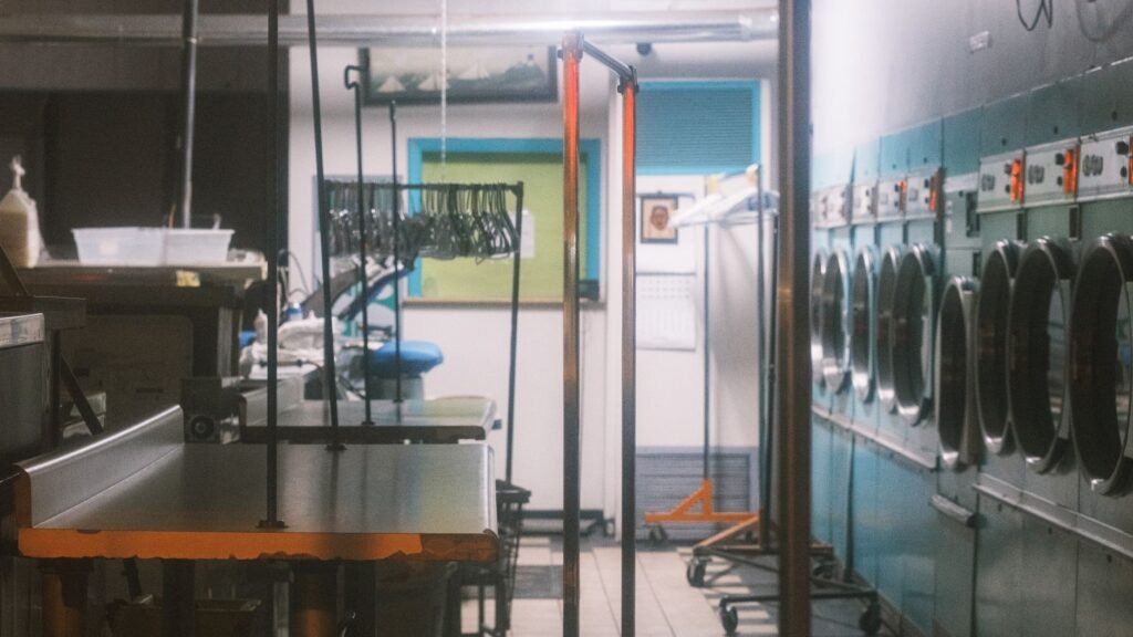 A dim commercial laundry with a row of pale blue tumble dryers down one wall, each control head showing small orange indicator lights